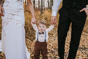 A toddler in brown suspenders holds hands with bride and groom parents in a fall forest setting. Wedding photography.