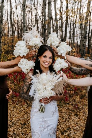 Smiling bride in lace gown surrounded by bridesmaids holding white bouquets in a fall forest. Wedding photography.