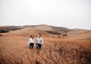 Couple walks hand in hand through golden autumn fields with rolling hills in the background. Couple photography. Engagement photos.