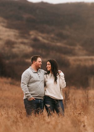 Couple standing together in an autumn field, gazing at each other with hills in the background. Couple photography. Engagement photos.