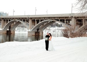 Bride and groom posing by snowy riverbank under arched bridge in winter. Wedding photography.