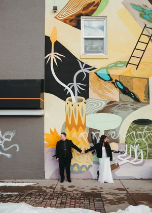 Couple holding hands in front of a colorful urban mural in winter, bride in white gown and groom in black suit. Wedding photography.
