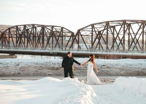 Bride and groom holding hands walking through snow with a historic iron bridge backdrop on their winter wedding day. Wedding photography.