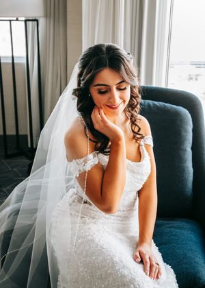 Bride in white lace gown and veil sitting by window on blue chair. Wedding photography.