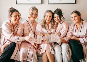 Five bridesmaids in pink satin robes holding drinks and smiling together. Wedding photography.