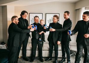 Groomsmen in black suits toasting with cans around small child indoors. Wedding photography.