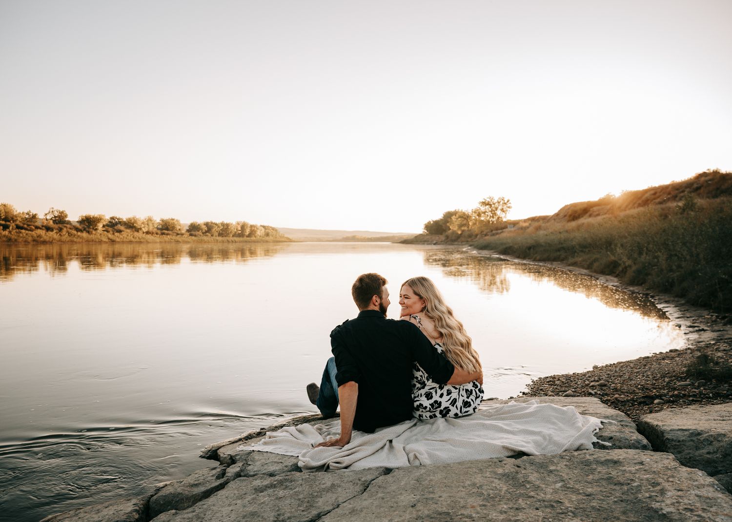 Couple sitting together on a blanket by a calm river at sunset, enjoying a peaceful romantic moment outdoors. Engagement photos.