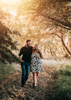 Couple walking together on a sunlit forest path, woman in floral dress and man in black shirt during golden hour. Engagement photos.