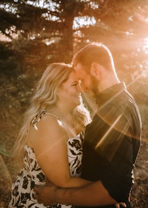 Couple sharing a romantic forehead-to-forehead moment bathed in warm golden sunlight among trees at sunset. Engagement photos.