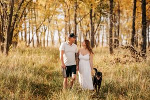 Couple walks with black dog through golden autumn meadow surrounded by yellow-leafed trees. Engagement photo session. Engagement photos.