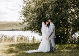 Bride and groom sharing a romantic kiss under a willow tree by a serene lake on their wedding day.