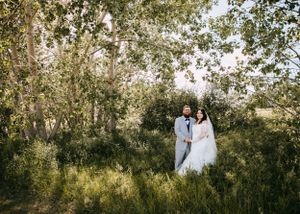 Bride and groom standing together surrounded by lush green trees and tall grass outdoors.