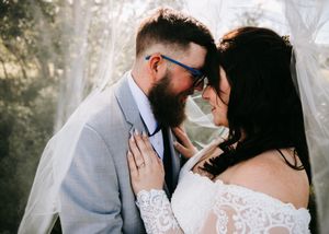 Bearded groom in grey suit and bride in lace off-shoulder gown sharing an intimate moment under a veil.