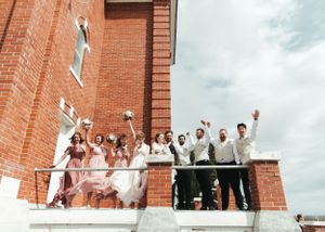 Wedding party celebrates on a brick church balcony, tossing bouquet into the sky.