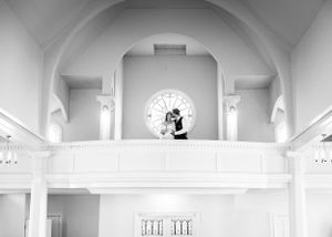 Black and white photo of bride and groom embracing on a church balcony with round window.