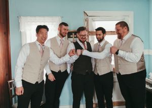 Groom and four groomsmen in tan vests laughing together during wedding preparations indoors.