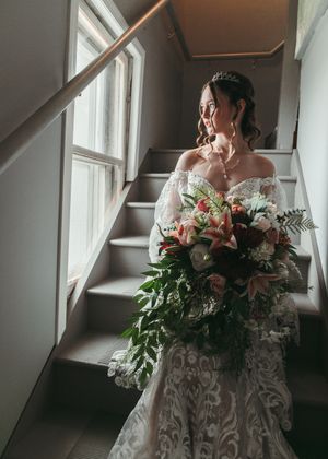 Bride in lace off-shoulder gown holding lush floral bouquet by staircase window.