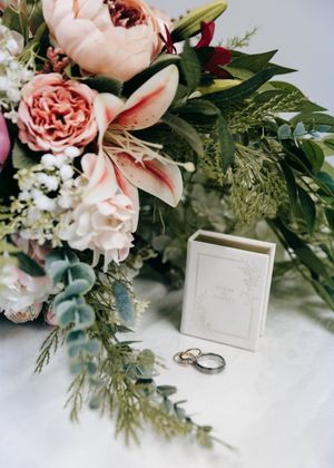 Wedding bouquet with pink peonies and lilies beside ring box and gold wedding bands.