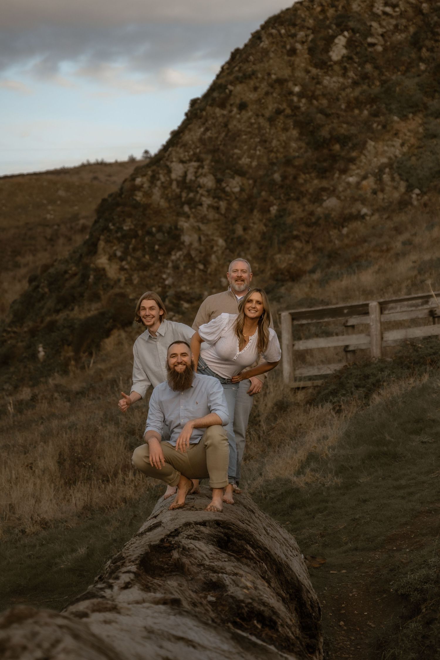 A group poses on a fallen log against a mountainous coastal backdrop at sunset.
