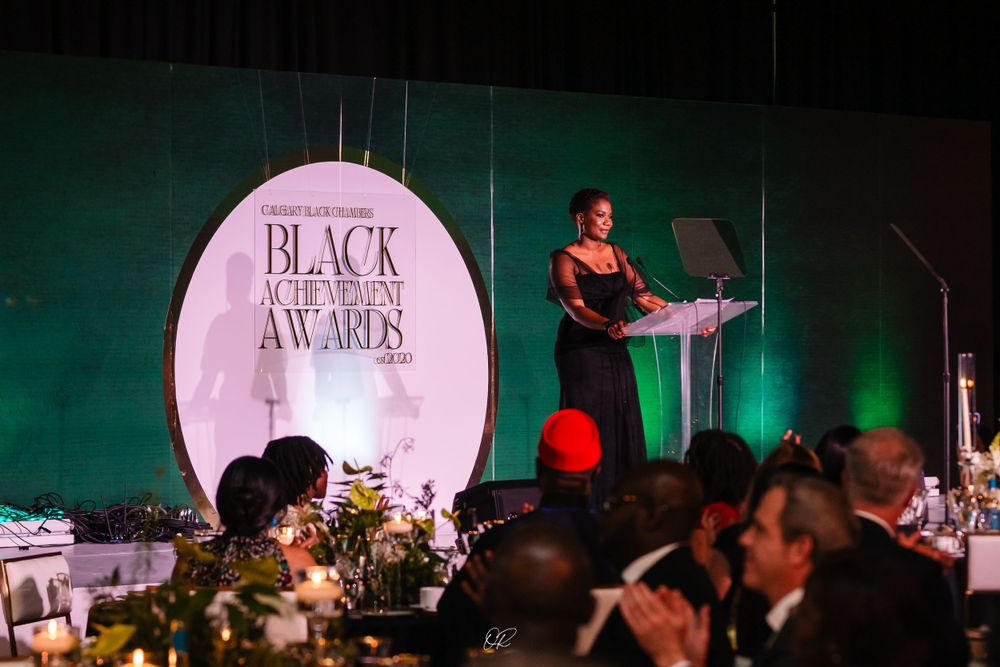 An awards ceremony podium with a speaker at a Black Excellence Awards event, with decorated tables and attendees visible.