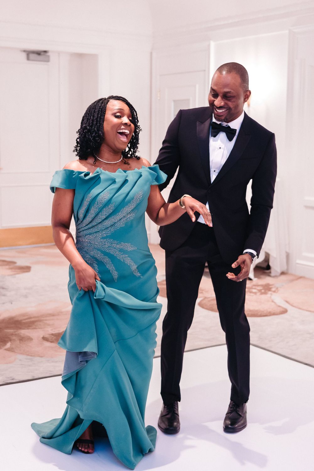 A couple shares a joyful dance moment at a formal event, with her wearing a turquoise gown and him in a black tuxedo.
