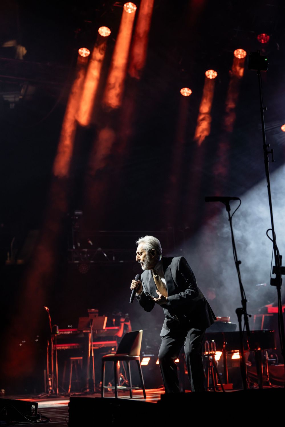 Dramatic spotlit performance on dark stage with orange lighting and atmospheric smoke effects.