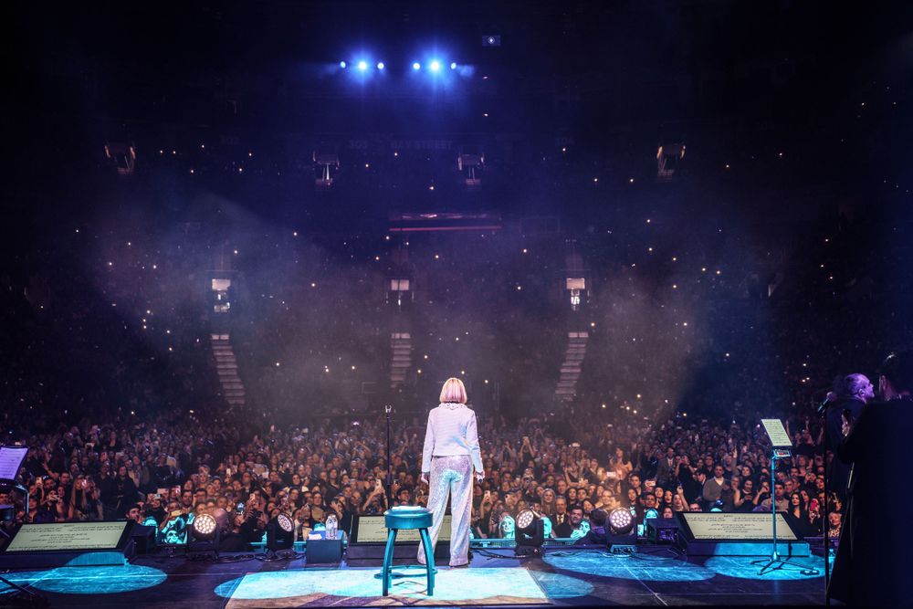 Wide arena concert shot with blue stage lighting and packed audience.