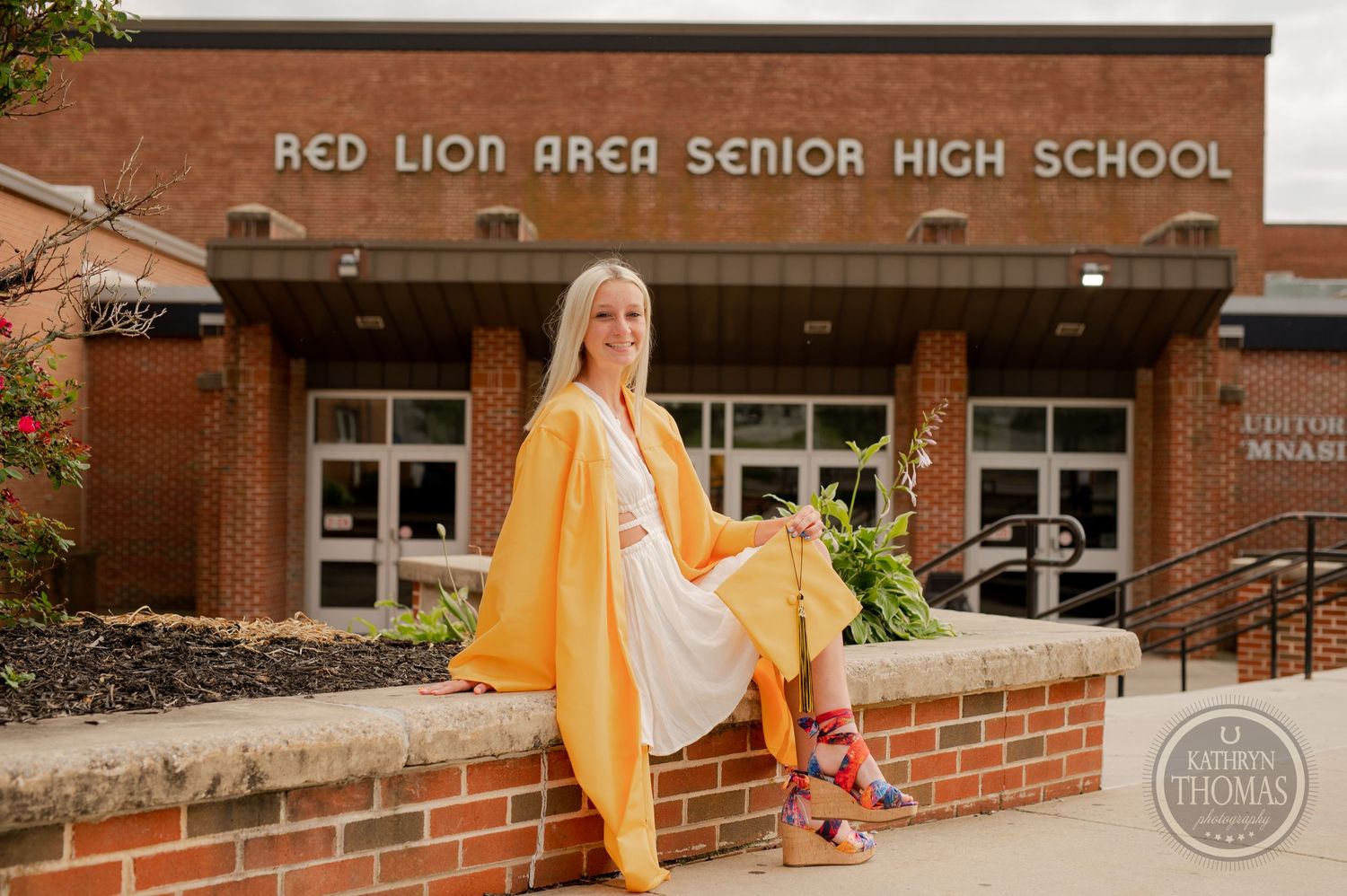 Graduate wearing bright yellow gown and colorful shoes poses on brick ledge at school entrance.