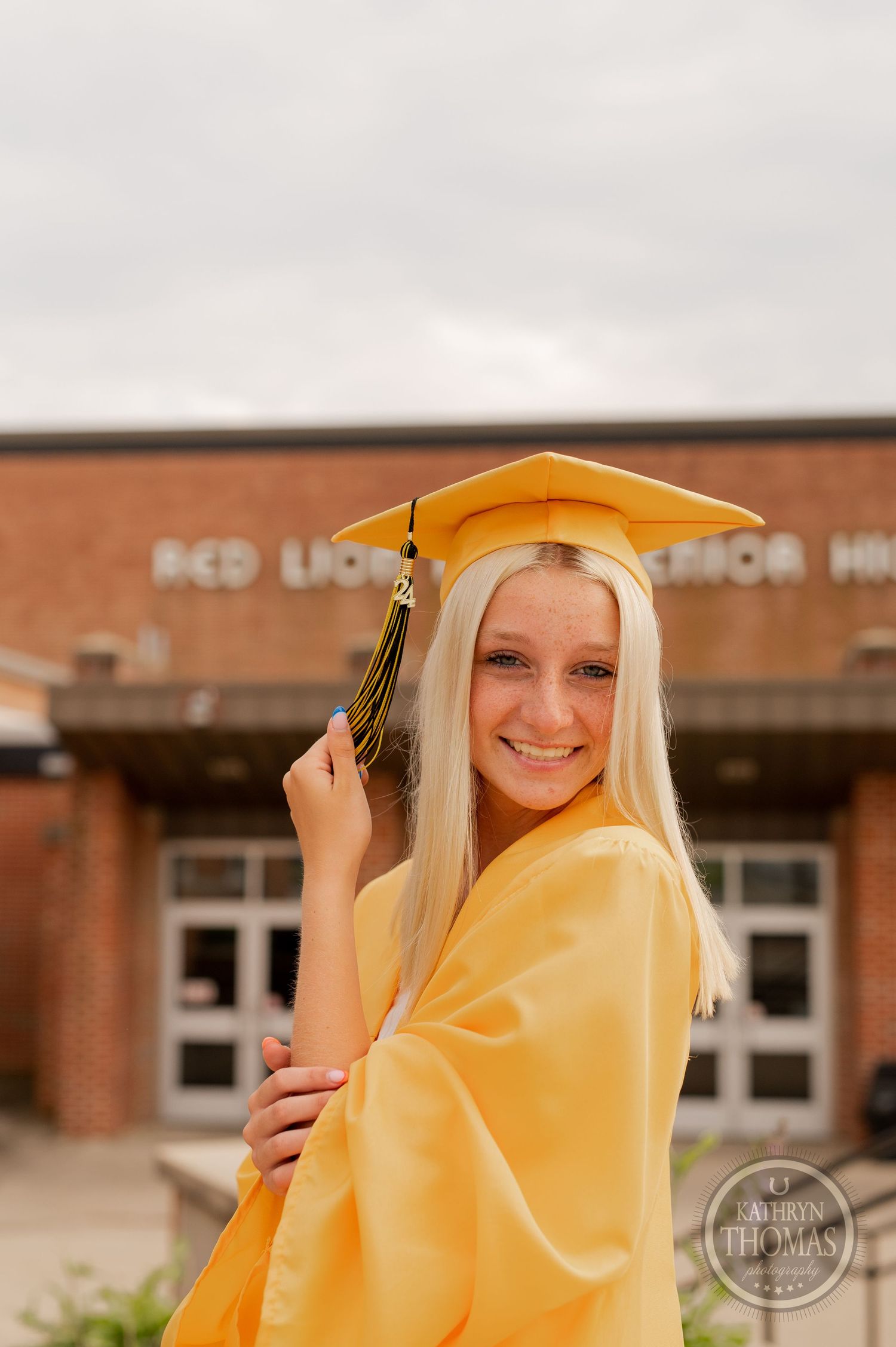Graduate in yellow graduation attire smiles while adjusting cap tassel outside school entrance.