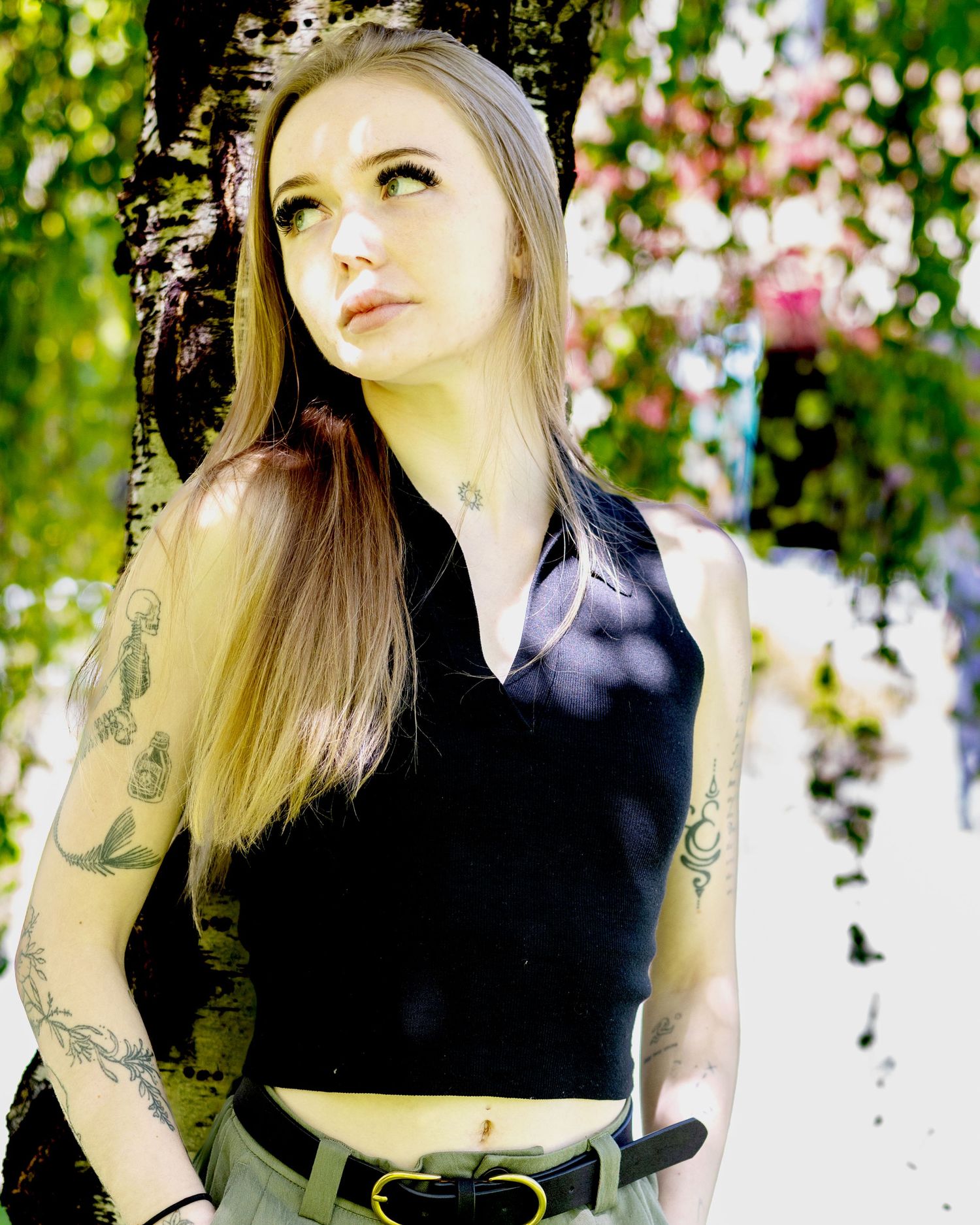 Gracie is posing under a tree with her back against the trunk, wearing a black V-neck top with no shoulders. You can see her tattoos on her-