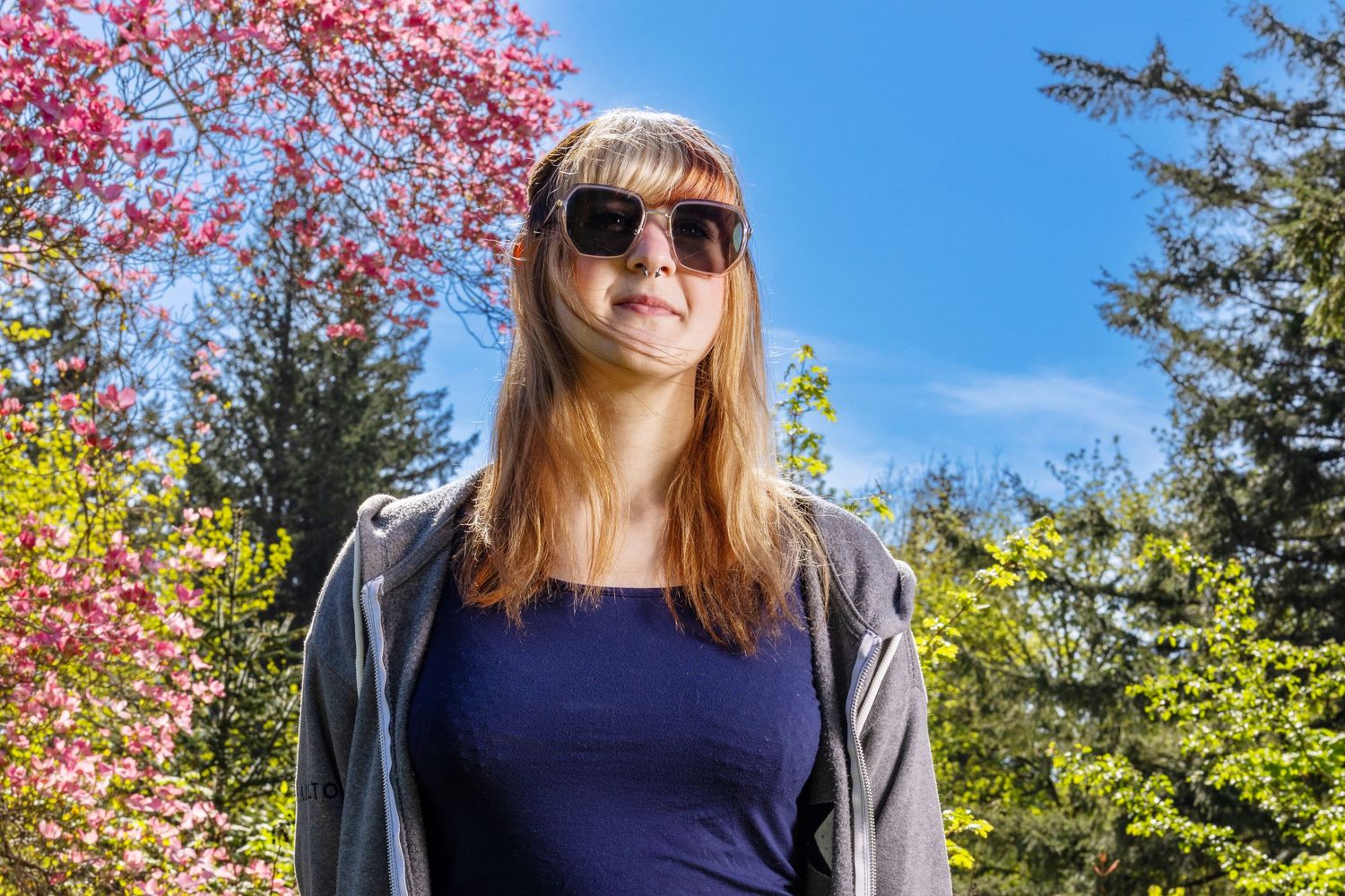 Izzy is wearing a dark sunglasses with a light gray jacket over her shoulders unzipped wearing a blue top. She is standing in front of green