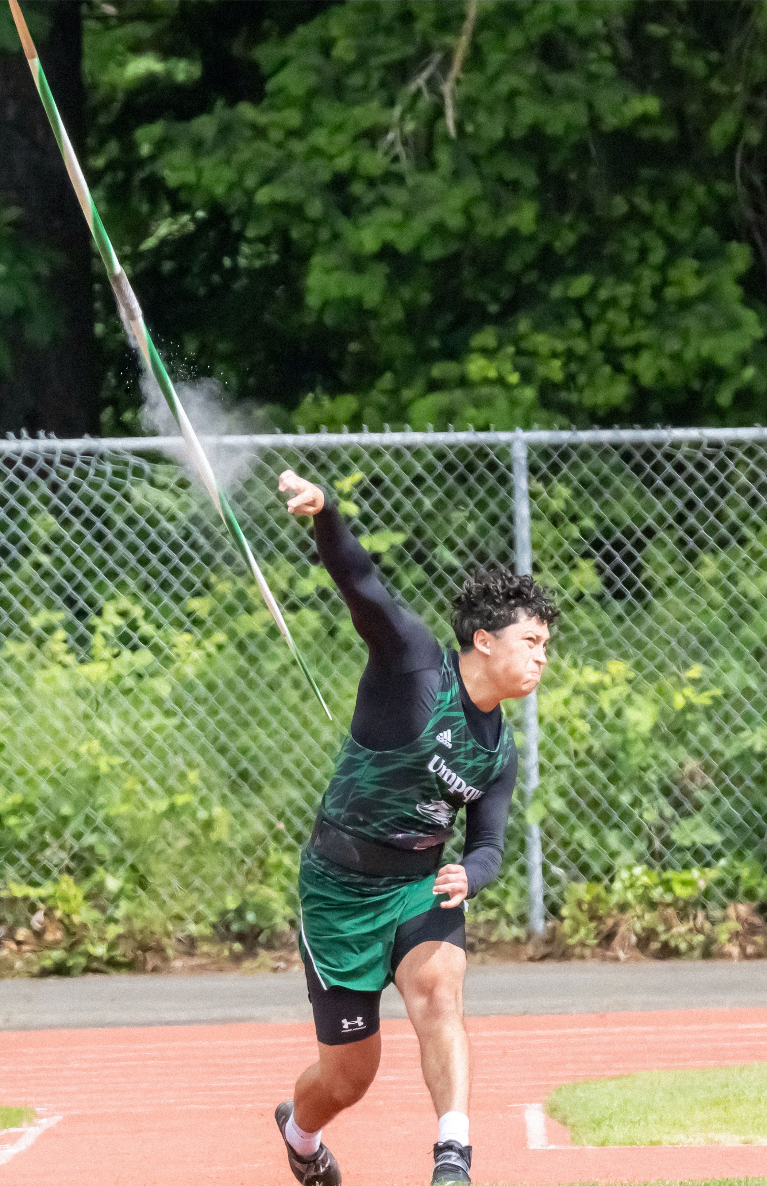 Track athlete releases a javelin on an outdoor field with trees and a chain-link fence behind.