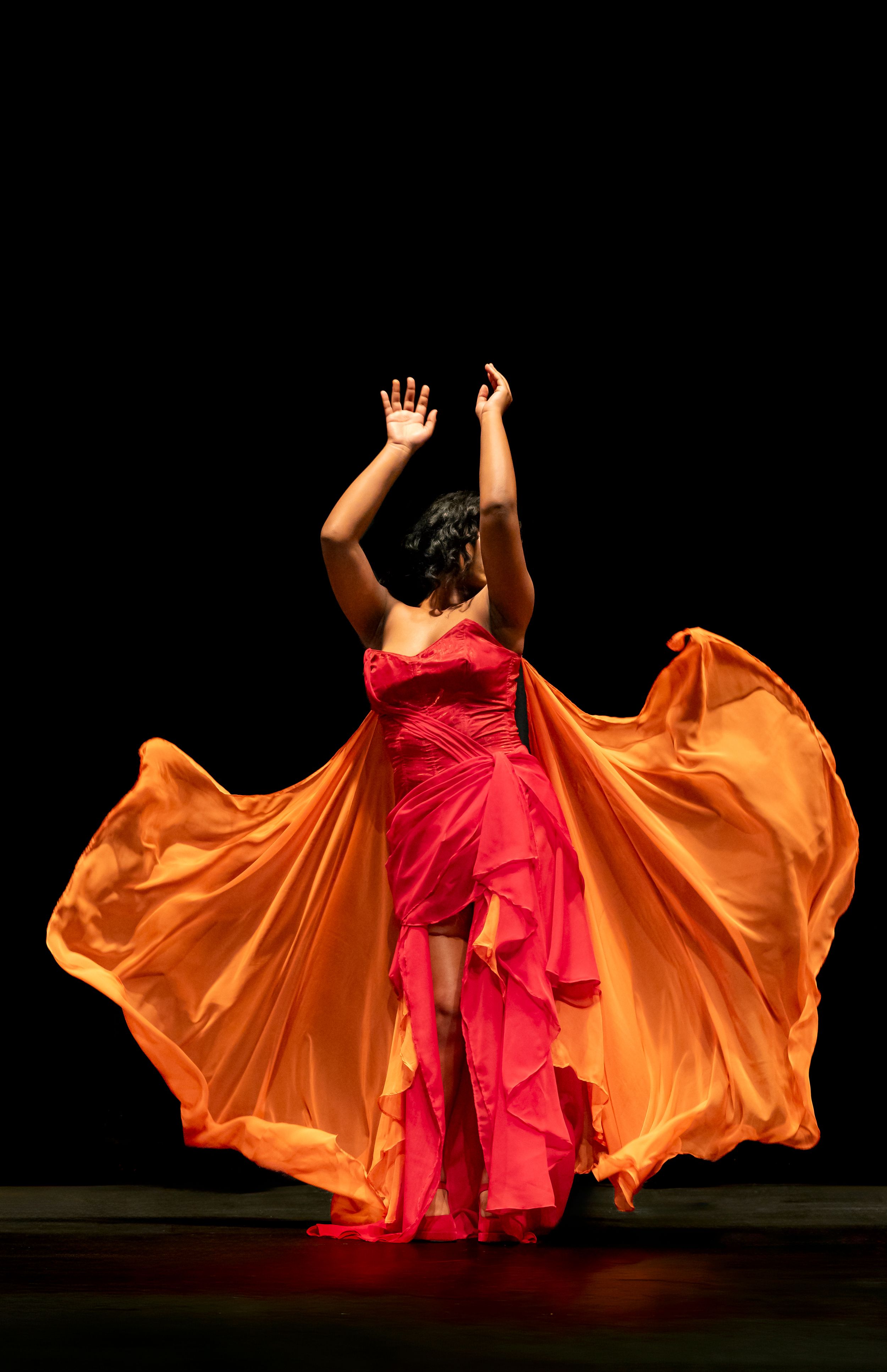 Dancer in a red dress raises her arms as orange fabric flows around her on a dark stage.