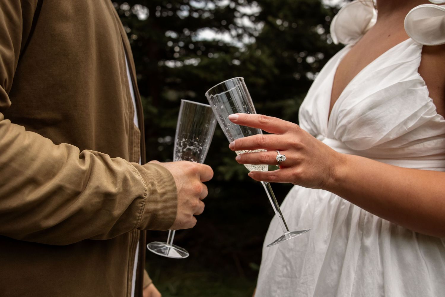 Close-up of two people toasting with champagne glasses outdoors, wearing brown jacket and white dress with diamond engagement ring.