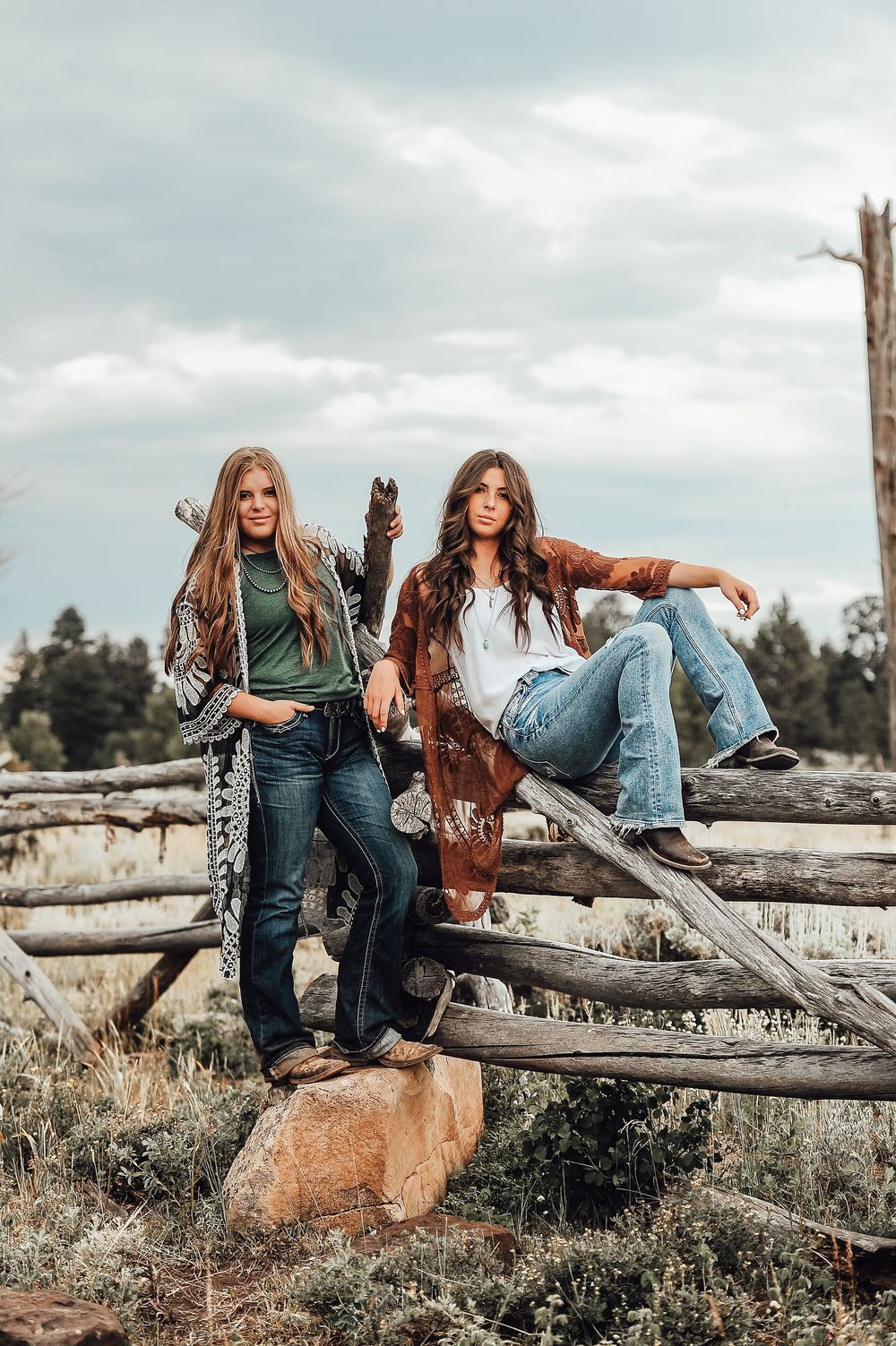 Two friends in casual wear posing on rustic wooden logs in a natural outdoor setting with a dog.