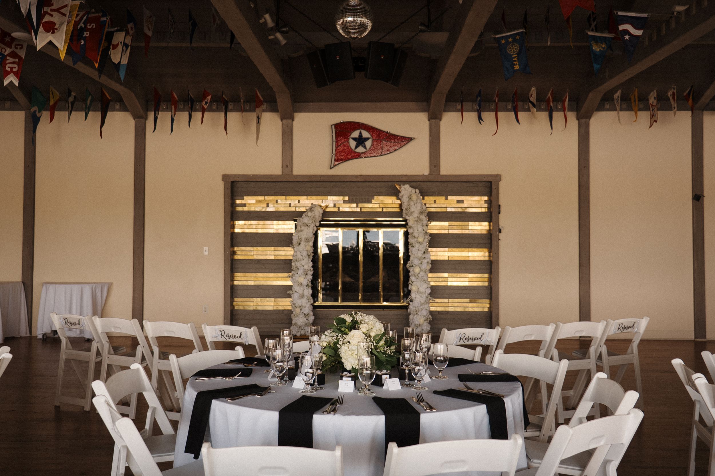 Modern ballroom with round dining tables, white chairs, and rustic wood wall panels lit by elegant chandeliers.