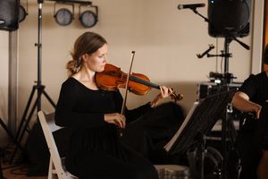 Professional musician performs on violin in a dark studio setting with lighting equipment visible in background.