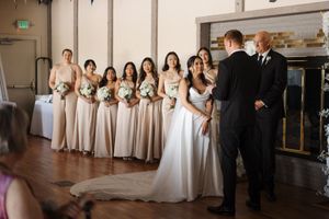 Wedding party members line up in formation wearing black suits and white dresses at an indoor venue.