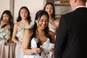Bride smiles joyfully while reading wedding vows during ceremony with bridesmaids in background.