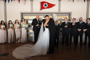 Wedding party poses for a formal group photo with bridesmaids in white and groomsmen in black against a wall.