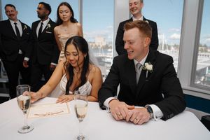 Group of wedding guests in formal black attire gathered around tables at a reception venue with city views.