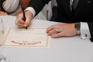 A close-up view of hands signing a document, with a formal paper and printed text visible in the frame.