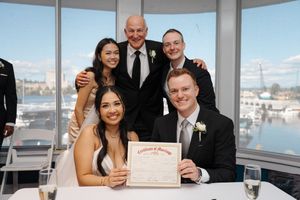 A group wearing formal attire poses together at a waterfront venue with a marriage certificate on the table.