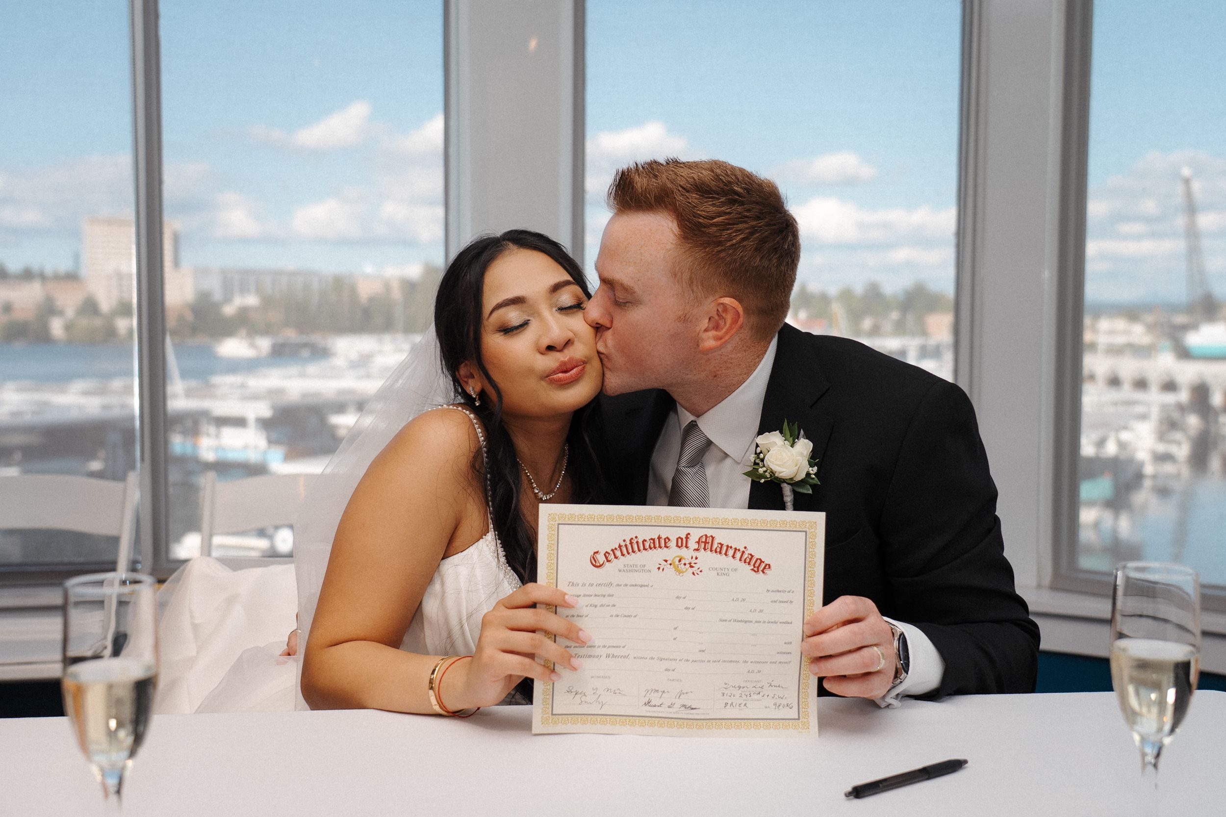 Couple kisses while sitting at a table with city views visible through floor-to-ceiling windows in an elegant setting.