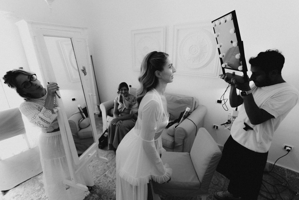 A wedding party in a white room gets ready before the ceremony, with a mirror being held up in the background.