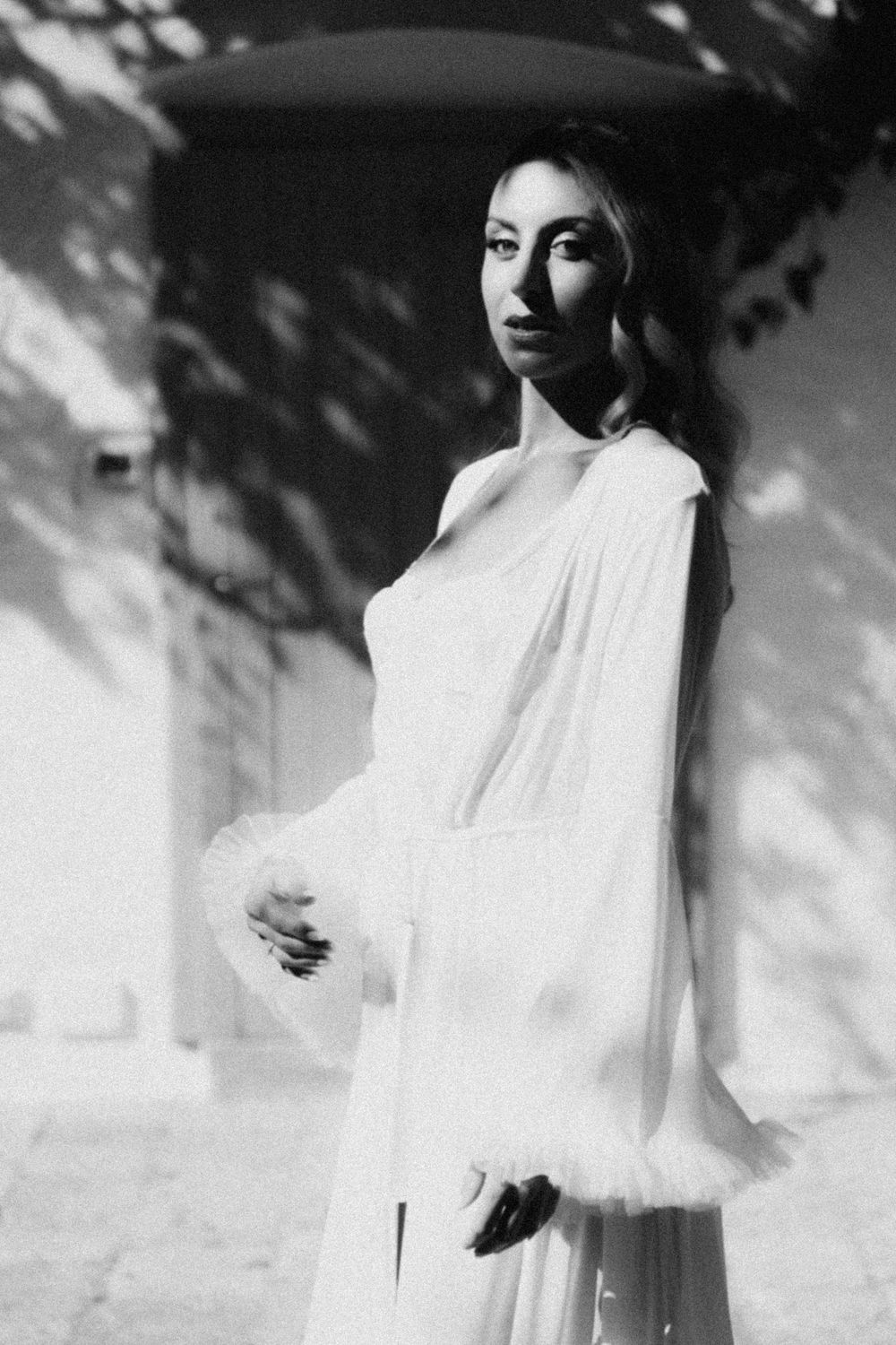 Dramatic black and white portrait of someone in a flowing white dress against a wall with dappled shadows.