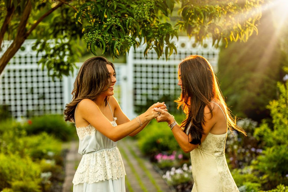 Two friends in white dresses hold hands joyfully in a sunlit garden setting with lattice backdrop.
