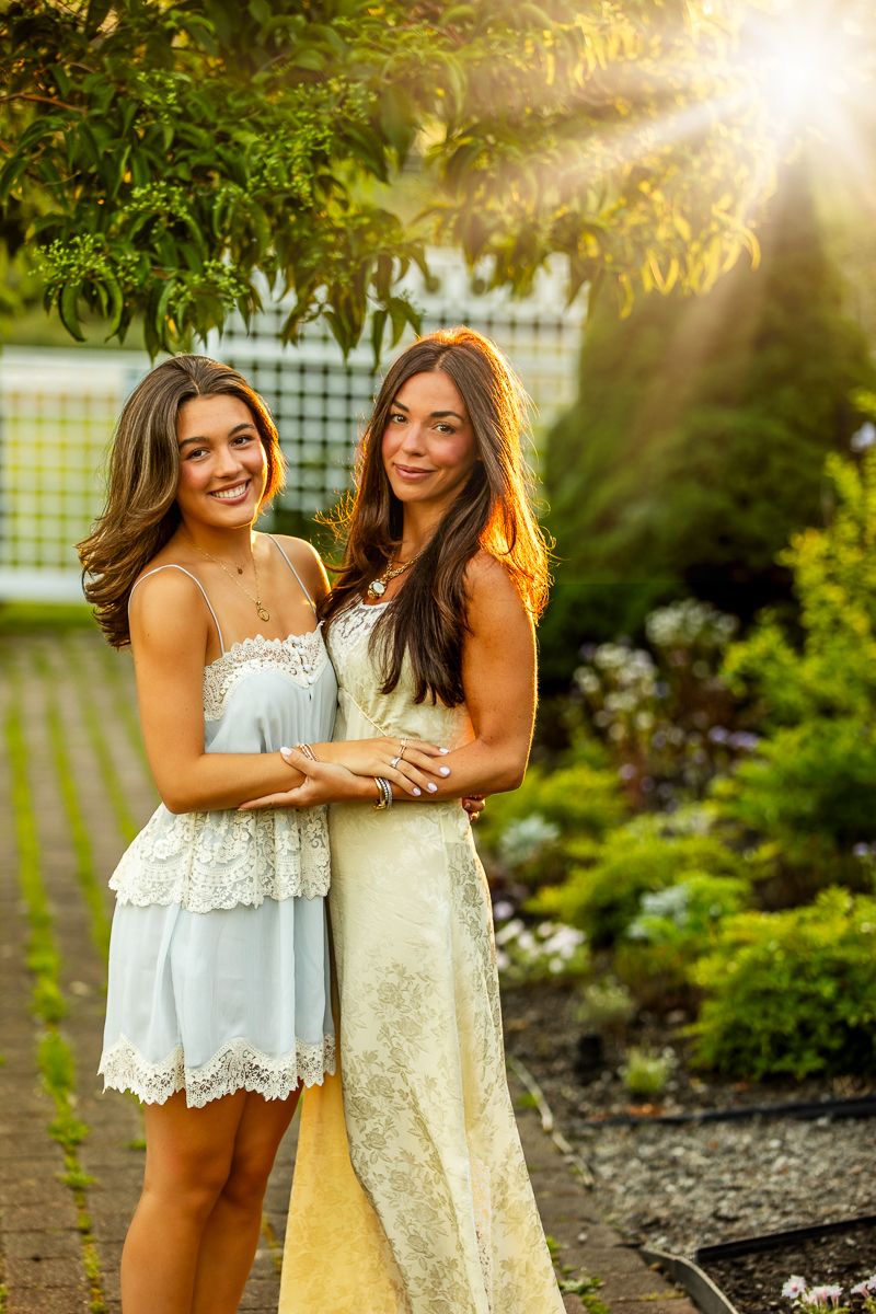 Two friends in summer dresses embrace and laugh together in a sunlit garden setting with lush greenery.
