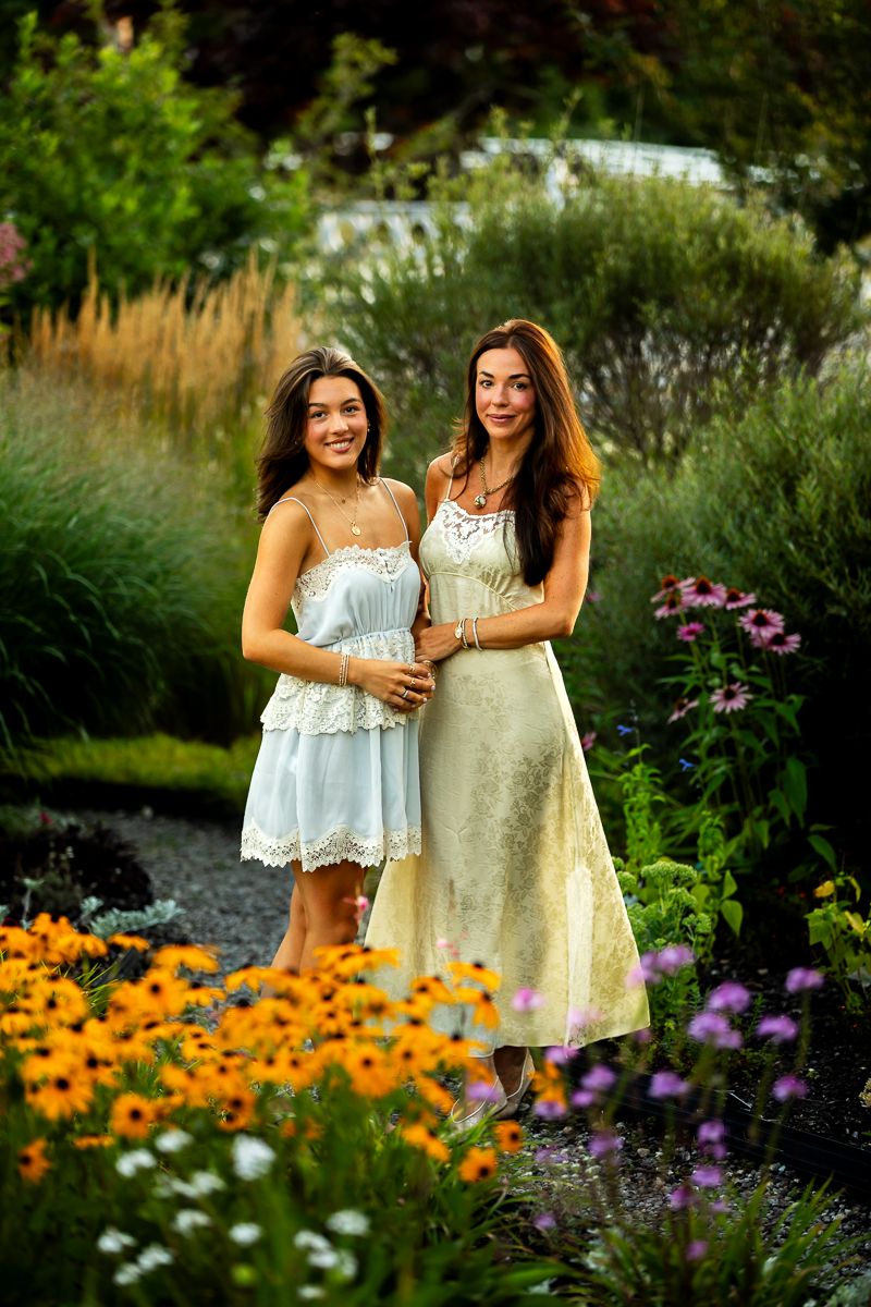 Two women in summery white dresses pose together in a vibrant garden with orange and purple flowers.
