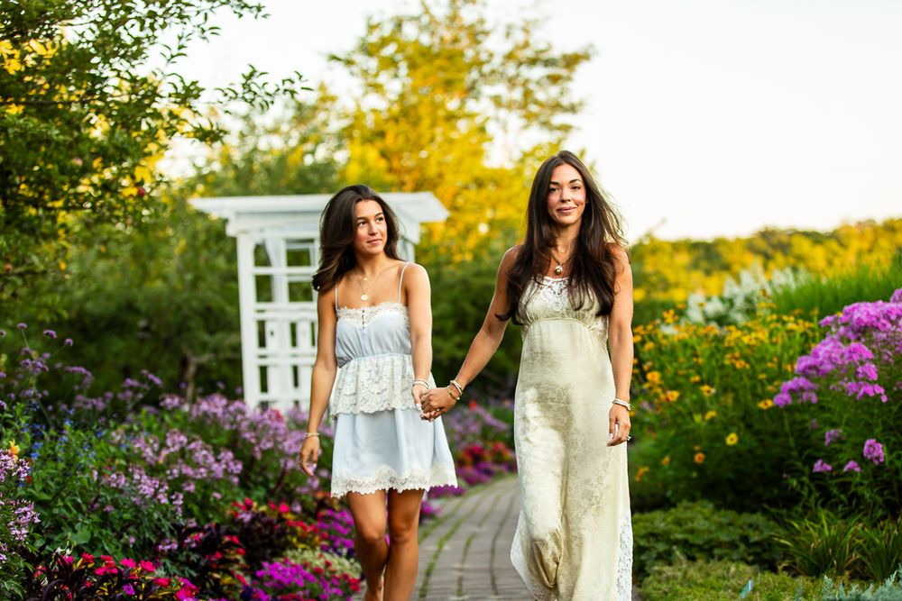 Friends in light dresses walk hand-in-hand through a colorful summer garden path at sunset.